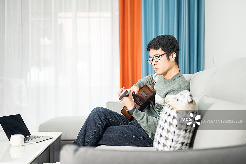 一位年轻的亚洲男子坐在家里的沙发上弹吉他图片素材