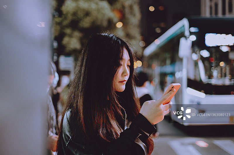 年轻亚洲女性在夜晚繁忙的街道上使用智能手机图片素材