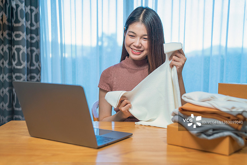 亚洲美女在家直播销售布料产品。年轻女子使用手机视频通话向顾客展示商品并介绍细节。远程购买和采购购物概念图片素材