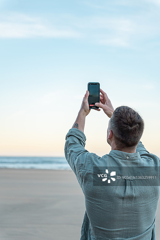 在海滩用手机拍摄日出的休闲蓝衣男人图片素材