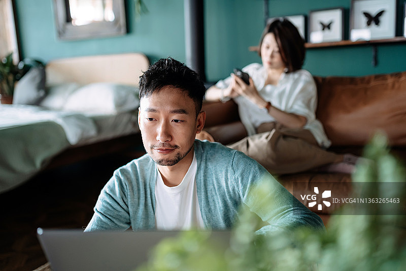 年轻的亚洲情侣坐在家里的客厅里，年轻女子在沙发上使用智能手机，而年轻男子坐在地板上使用笔记本电脑。日常生活中的科技图片素材