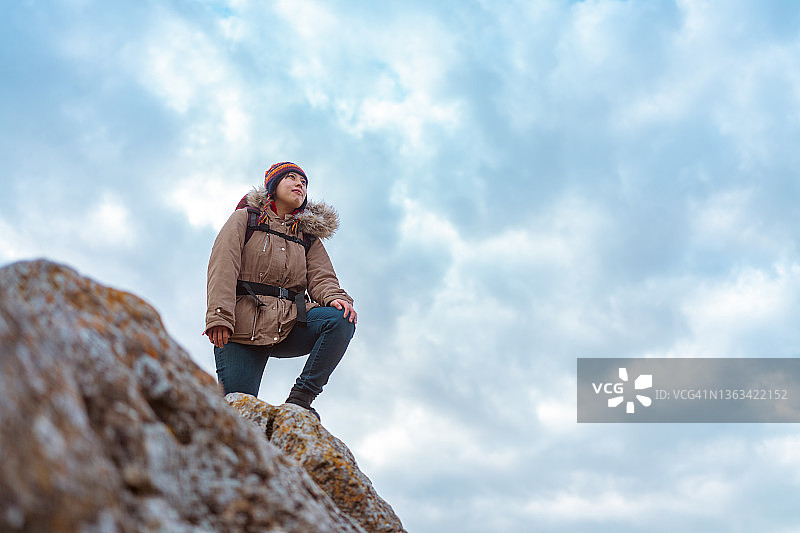 年轻女徒步者站在冬日多云天空下的山顶岩石上图片素材