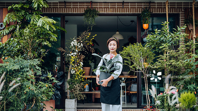 自信的亚洲女花店主，站在花店外，手捧盆栽，面带微笑图片素材