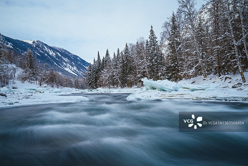 森林中被白雪覆盖的树木旁的冰冻河流图片素材