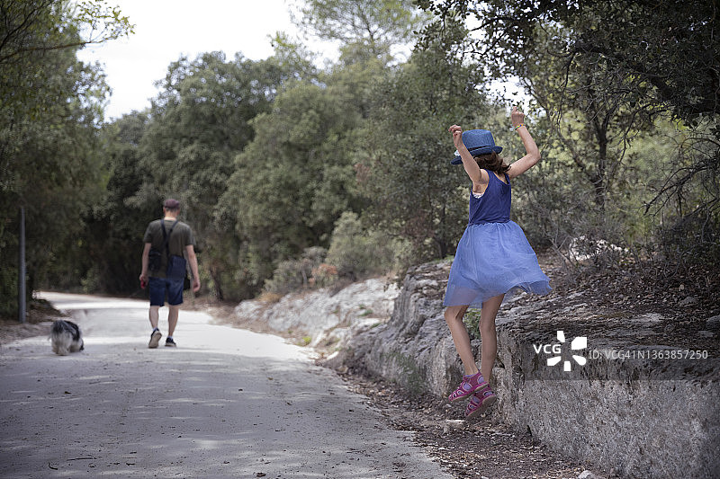 小孩（8-9岁）在乡村小路上与父亲和宠物狗同行，跳下石墙图片素材