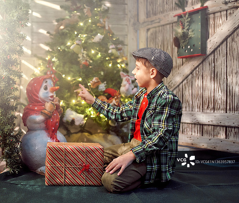快乐的男孩坐在圣诞树旁，指着玩具雪人图片素材