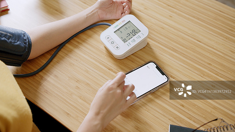 亚洲女人在家用智能手机记录血压监测仪上的血压和心率数据图片素材