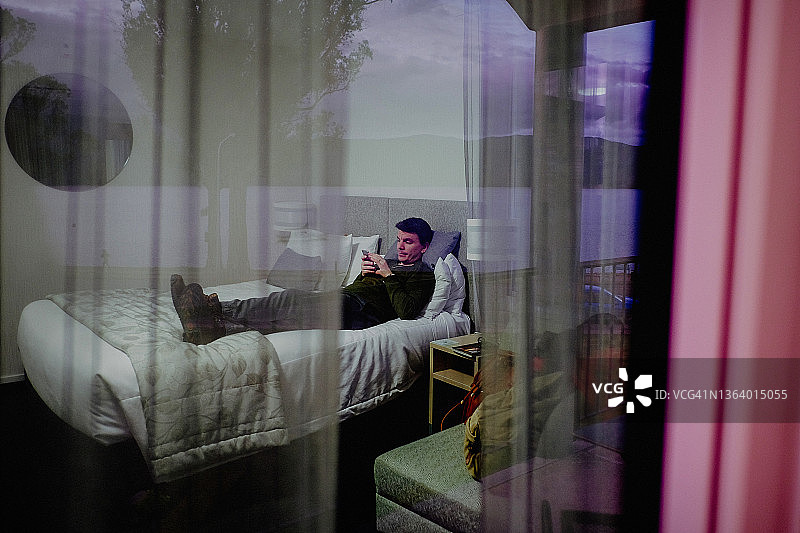 中年男人躺在床上看手机，透过窗户看风景图片素材