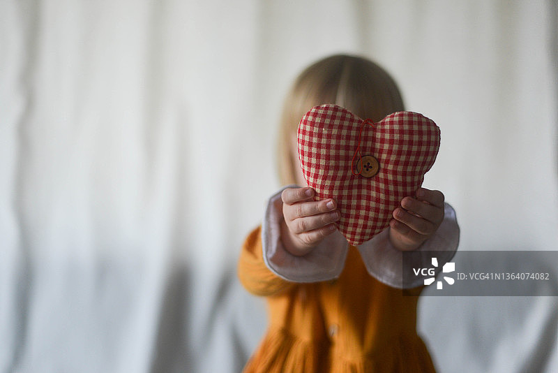 穿着橙色连衣裙的小女孩坐在蓝色沙发上，手捧自制爱心，祝爸爸或朋友情人节快乐。无脸图片素材