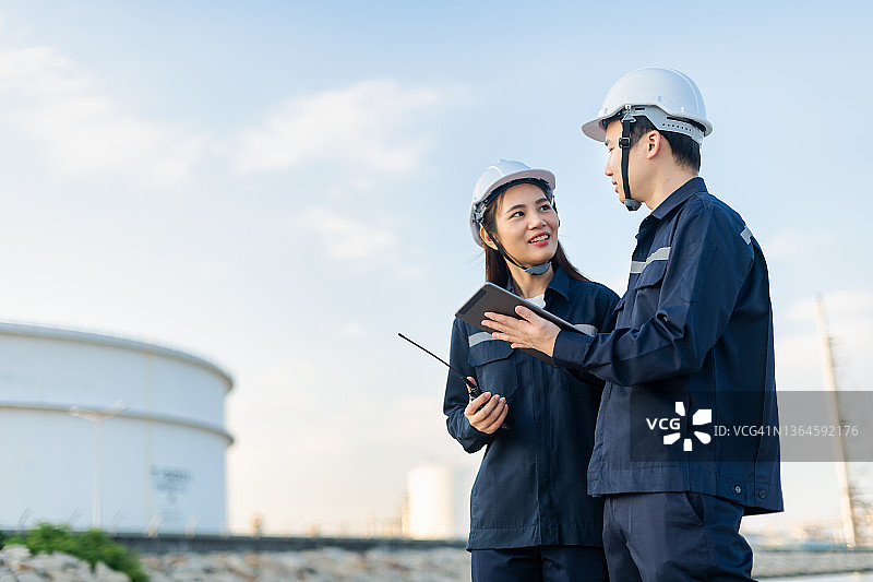 石油和天然气工业中佩戴个人安全防护设备（PPE）的炼油工程师，通过平板电脑与团队进行无线电通讯，检查并遵循清单。经典能源商业理念。图片素材