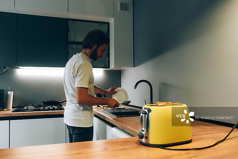 年轻男人在厨房洗碗。早晨，黄色烤面包机，留着胡子的男人，现代厨房，房屋清洁。图片素材