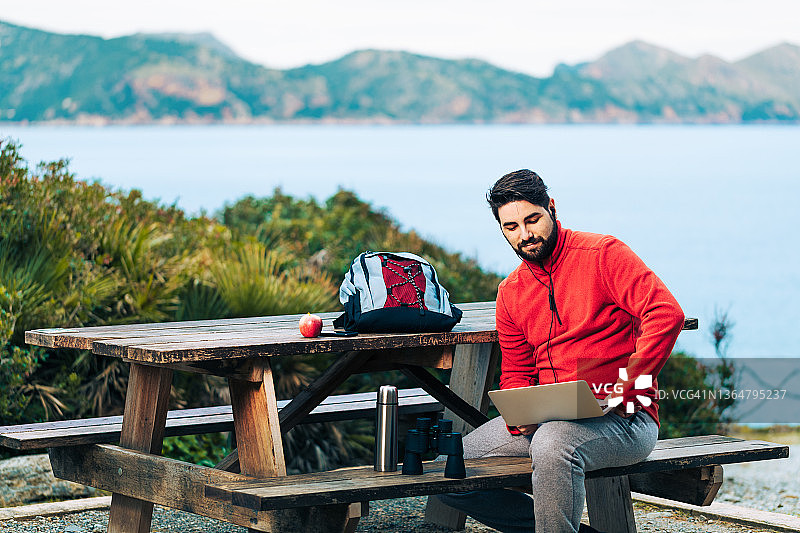年轻徒步者在露营地使用笔记本电脑和耳机，背景是马略卡岛北部海岸图片素材