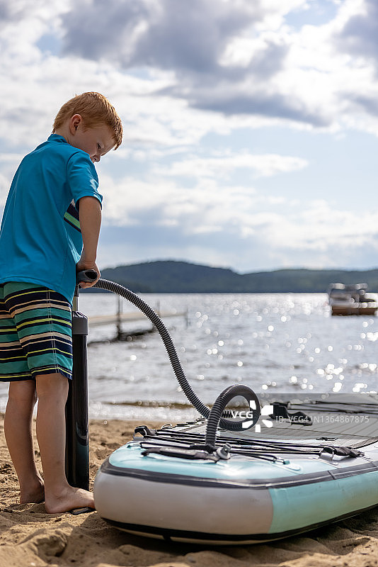 在魁北克夏季海滩给桨板SUP充气的可爱红发男孩图片素材