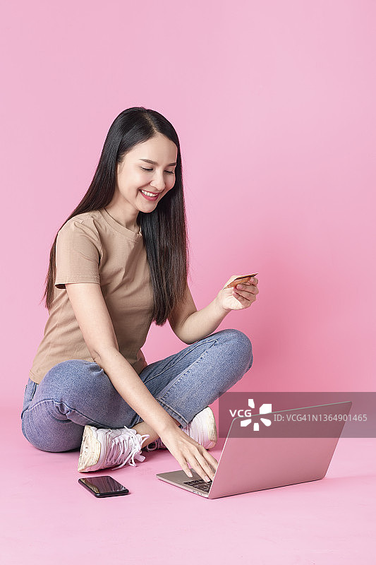 快乐的女人使用笔记本电脑和智能手机，拿着信用卡，对着粉红色背景微笑图片素材