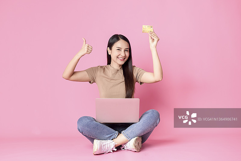 自信的年轻女子使用笔记本电脑，并对着粉色背景竖起大拇指，同时拿着信用卡图片素材