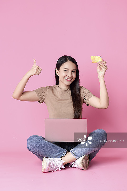自信的年轻女子使用笔记本电脑，并对着粉色背景竖起大拇指，同时拿着信用卡图片素材