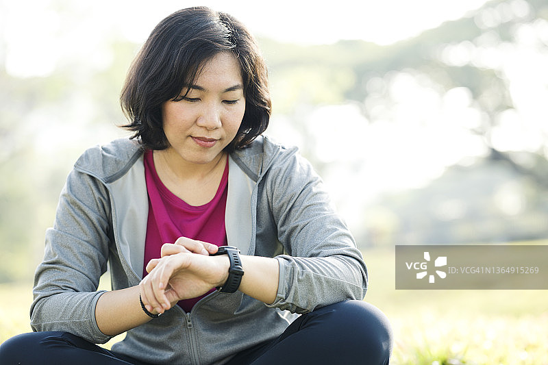 在户外运动时使用智能手表追踪心率。一位穿着运动服的亚洲女跑步者在慢跑后查看智能手表，以追踪她的步数和心率变化。图片素材