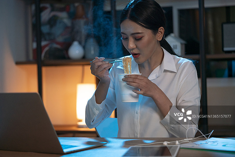 年轻女子坐在客厅里一边吃面条一边用电脑工作图片素材