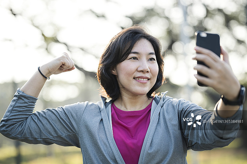 在运动中玩得开心做自己。亚洲女人在城市公园户外运动后，展示肌肉给视频通话中的朋友看图片素材