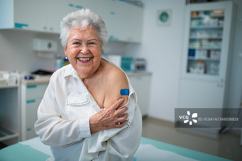 接种疫苗后手臂贴着绷带的快乐老年妇女在医生办公室看着镜头图片素材