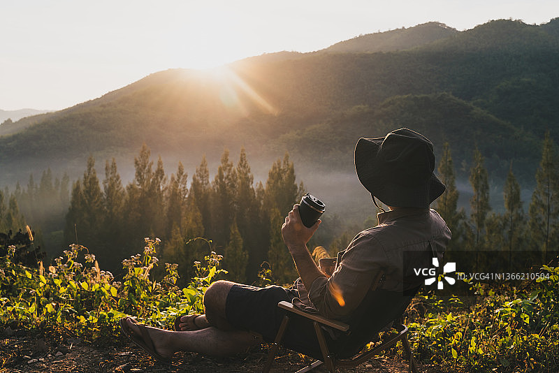 坐在露营椅上的男人在夏天享受露营，手持咖啡杯欣赏早晨的山景的背影图片素材