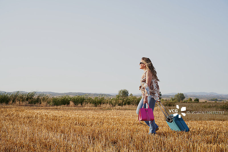 中年女人在稻草地上行走并拉着小型轮式行李箱，旅行概念图片素材
