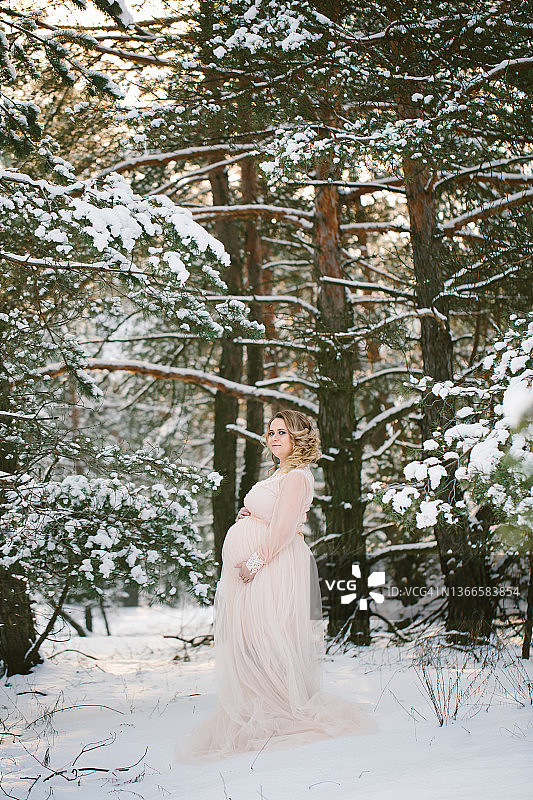怀孕的金发女人，发型和妆容精致，身穿复古长裙，站在冬季白雪皑皑的森林里，对着镜头微笑，用手托着怀孕的肚子图片素材