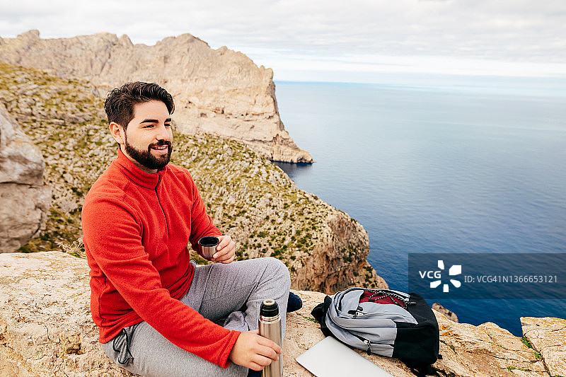 年轻徒步者坐在岩壁上，用笔记本电脑享用保温瓶里的热茶，背景是马略卡岛波连萨海湾的北部海岸图片素材
