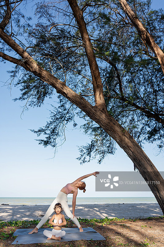 阳光明媚的夏日，亚洲母亲在海滩教女儿做瑜伽图片素材