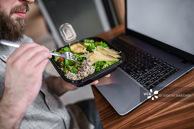 在笔记本电脑上做自由职业的络腮胡男子享用金枪鱼沙拉图片素材