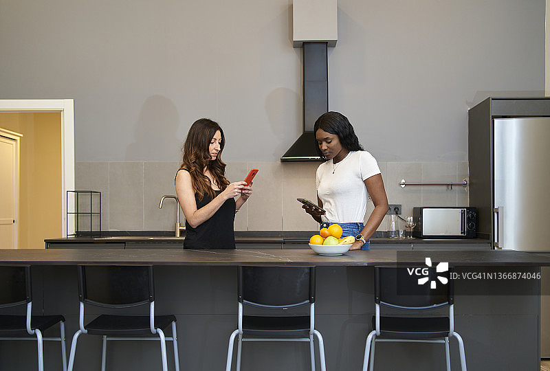 聚集在家中、站在厨房柜台后使用手机的女性朋友正面图图片素材