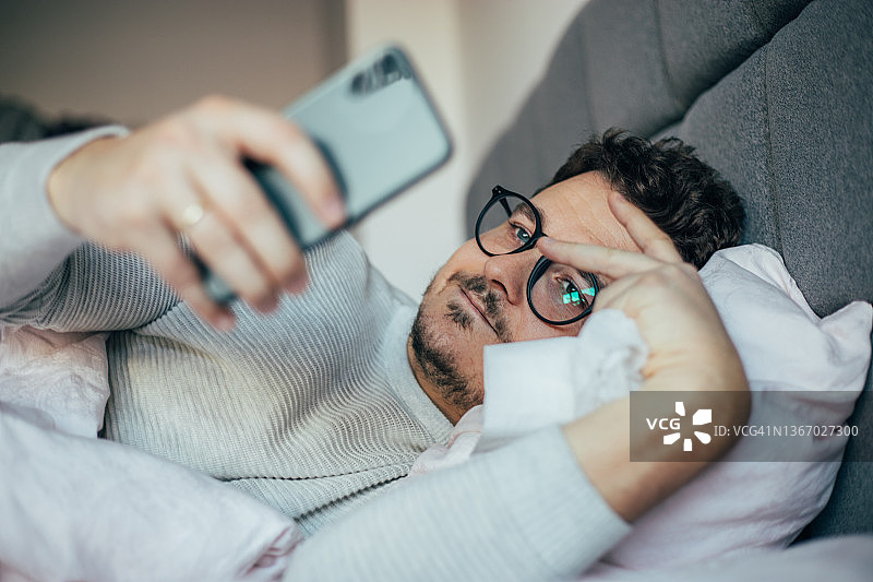生病的男人在床上休息时查看智能手机图片素材