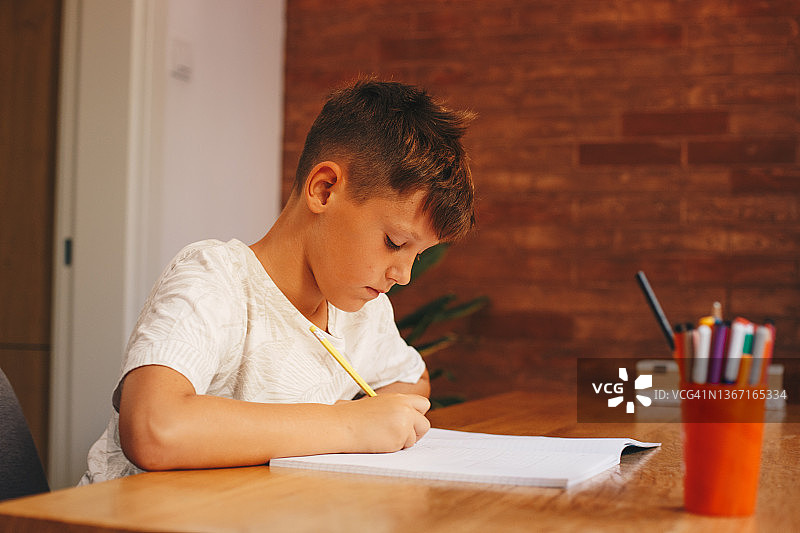 穿着白色T恤的男孩坐在桌旁写作业。图片素材