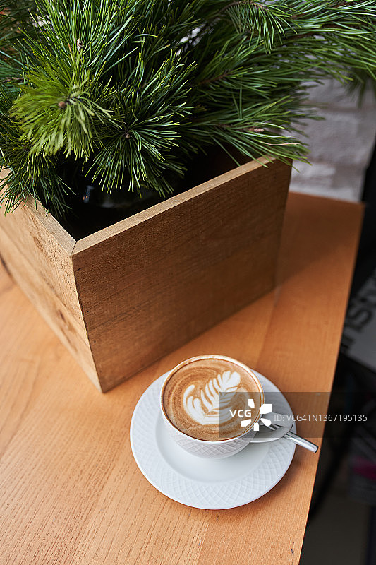 窗边木制窗台上的一杯带有拿铁拉花的白色咖啡，窗外可见松枝和烛台。图片素材