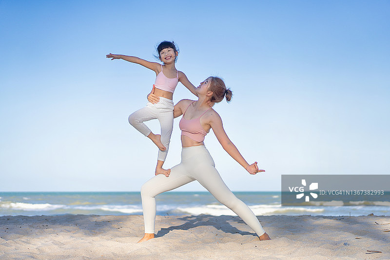 阳光明媚的夏日，亚洲母亲在海滩教女儿做瑜伽图片素材