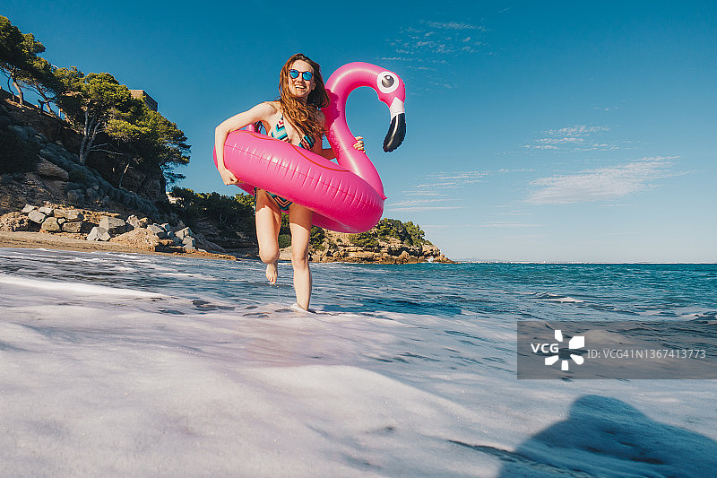 快乐的年轻女子在海滩上享受阳光明媚的一天，带着充气火烈鸟图片素材