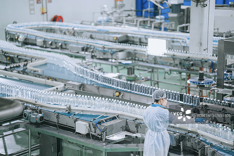 在饮用水工厂生产线上使用个人防护装备检查水瓶的亚洲华人男工人，进行日常工作图片素材