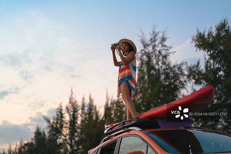 戴太阳帽站在车顶拍摄海滩日落景色的女人图片素材