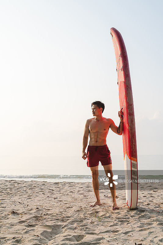 站在海滩上的亚洲年轻运动员与桨板的肖像图片素材