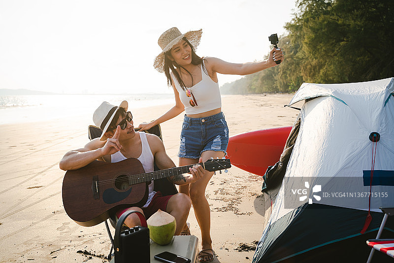 年轻的亚洲情侣在海滩上享受露营，一边弹吉他一边自拍。图片素材