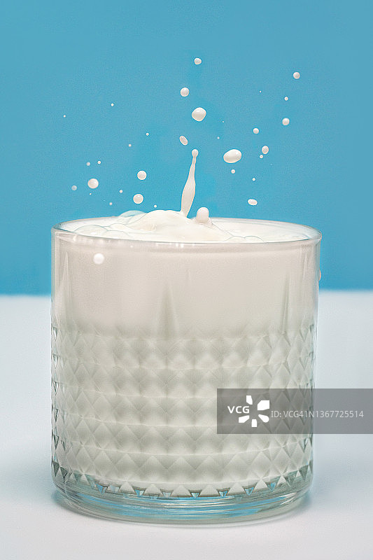 在柔和的蓝白色背景上，装有牛奶和飞溅物的透明玻璃杯图片素材
