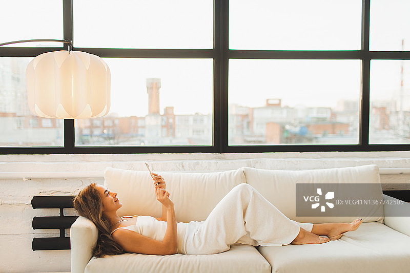 微笑的女人躺在沙发上玩智能手机，浪费时间上网，用小工具拖延时间图片素材