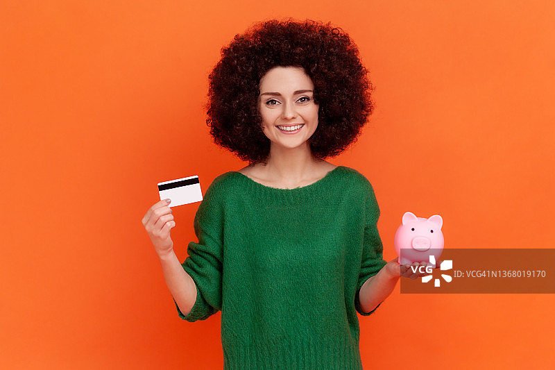 有着非洲发型的年轻女人穿着绿色休闲毛衣，对着镜头展示小猪存钱罐和信用卡图片素材