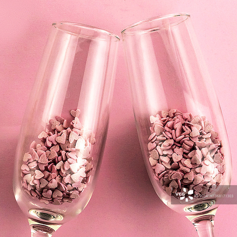两只香槟酒杯在粉红色背景上碰撞，带有红色心形彩纸图片素材