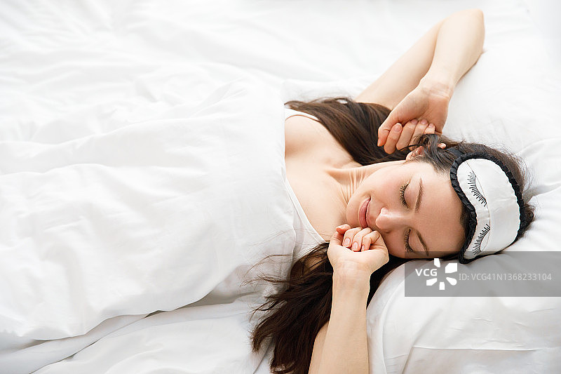 年轻女子从睡梦中快乐醒来。戴着睡眠面罩的女人在床上伸展身体，看着镜头微笑。美好的一天概念图片素材