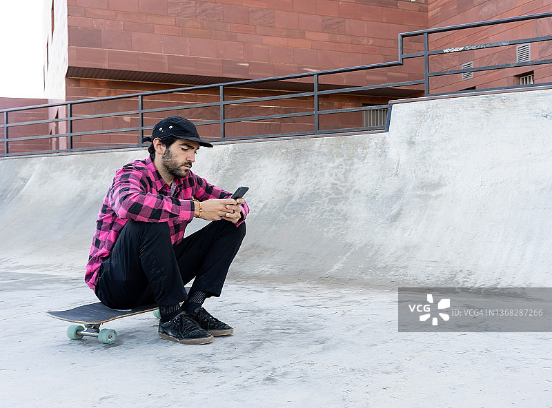 戴帽子在滑板公园玩手机的格子衬衫男人图片素材