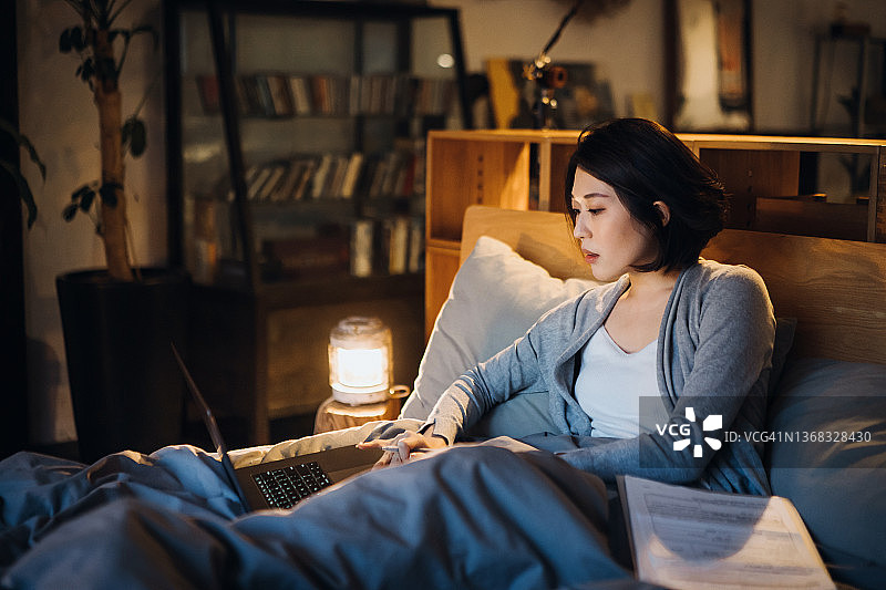 忙碌的年轻亚洲女人坐在床上，在家用笔记本电脑工作，晚上在家处理文书工作图片素材