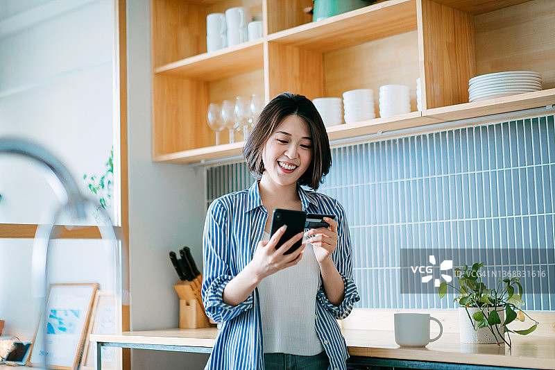 微笑的亚洲年轻女性在家中厨房用手机和信用卡管理账单支付图片素材