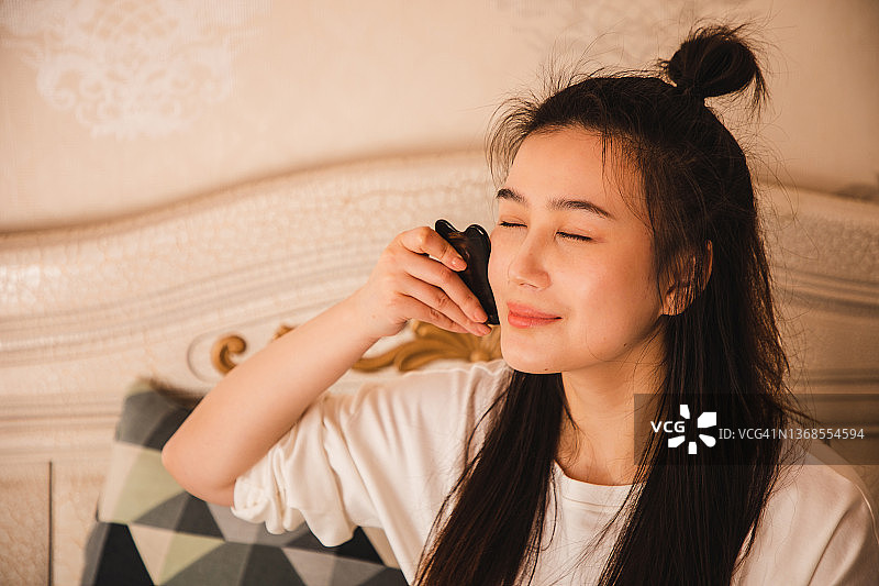 亚洲妇女在卧室里用刮痧板做面部按摩图片素材