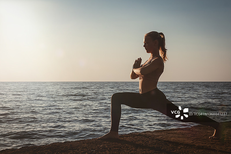 户外瑜伽：运动型身材女性在海边练习阿斯汤加串联瑜伽 Virabhadrasana 1 战士第一式图片素材